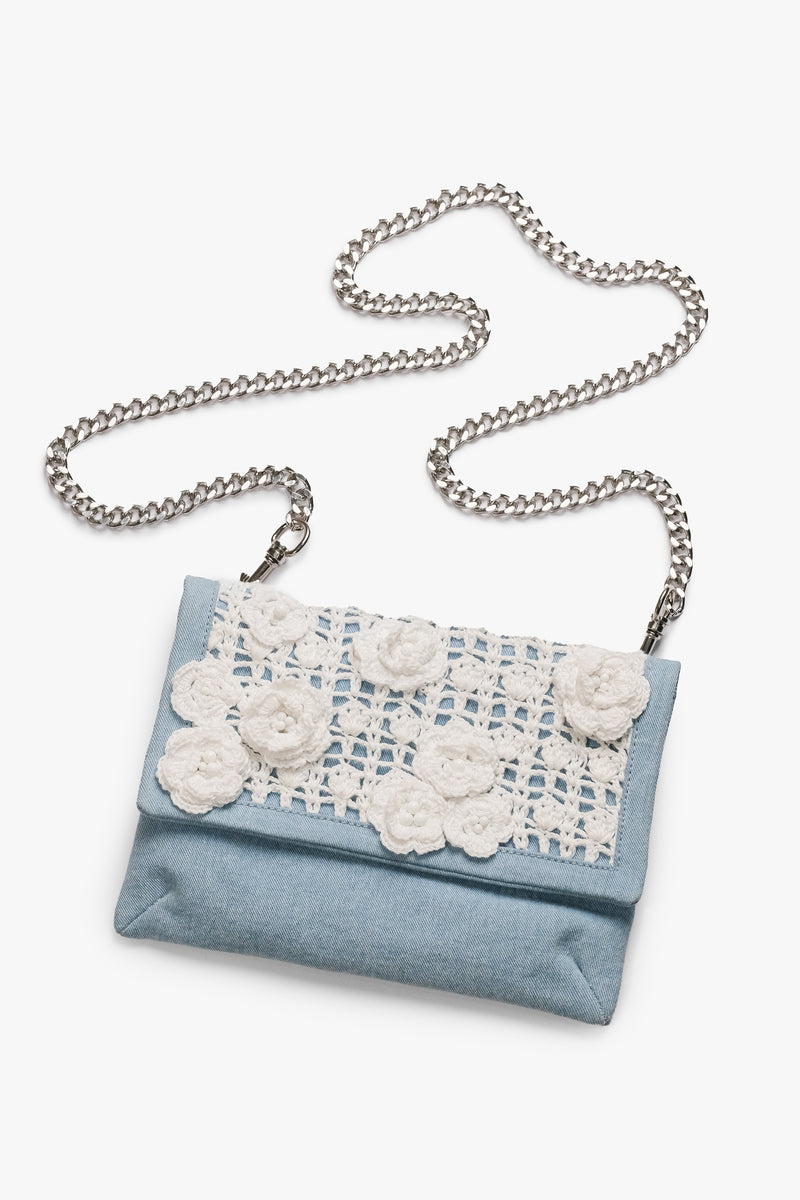 Acambha's Crochet Flower Clutch features a delicate crochet panel with flowers and bead detailing on light-colored denim. It has a zipper and magnetic flap closure, with a detachable chain strap for versatility